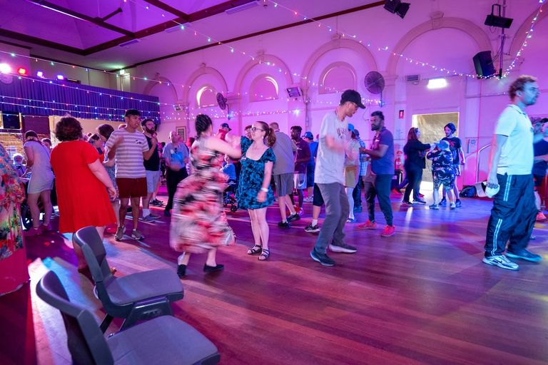 People dancing at the Rock the Coast event inside a brightly lit hall decorated with string lights and purple ambient stage lighting.